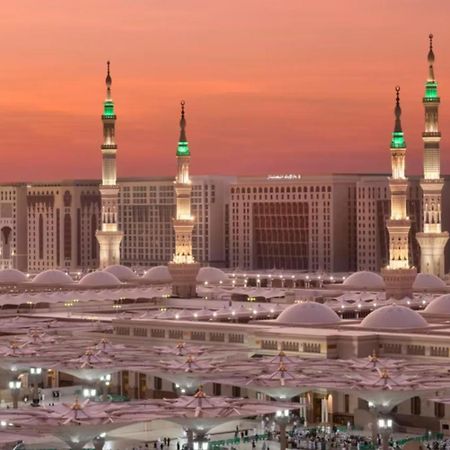 Hotel Intercontinental Dar Al Iman Madinah By Ihg Medina Exterior foto The photo depicts a scenic view of a mosque complex, likely during sunset, with several minarets illuminated and showcasing green lights. The architecture features domed roofs, and the setting includes a backdrop of buildings, all bathed in a warm, o