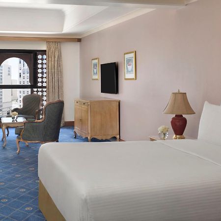 Hotel Intercontinental Dar Al Iman Madinah By Ihg Medina Exterior foto The photo shows a well-furnished hotel room. It features a large bed with a white duvet, two bedside lamps, and decorative paintings on the walls. There is a comfortable seating area with two chairs and a small table. A wooden dresser is positioned a