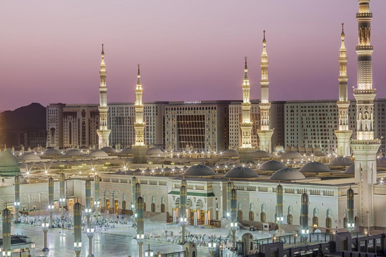 Hotel Intercontinental Dar Al Iman Madinah By Ihg Medina Exterior foto The photo shows a view of an expansive mosque complex, featuring several tall minarets that are illuminated against a twilight sky. The structure appears grand and is likely adorned with intricate architectural details. There are people gathered in t