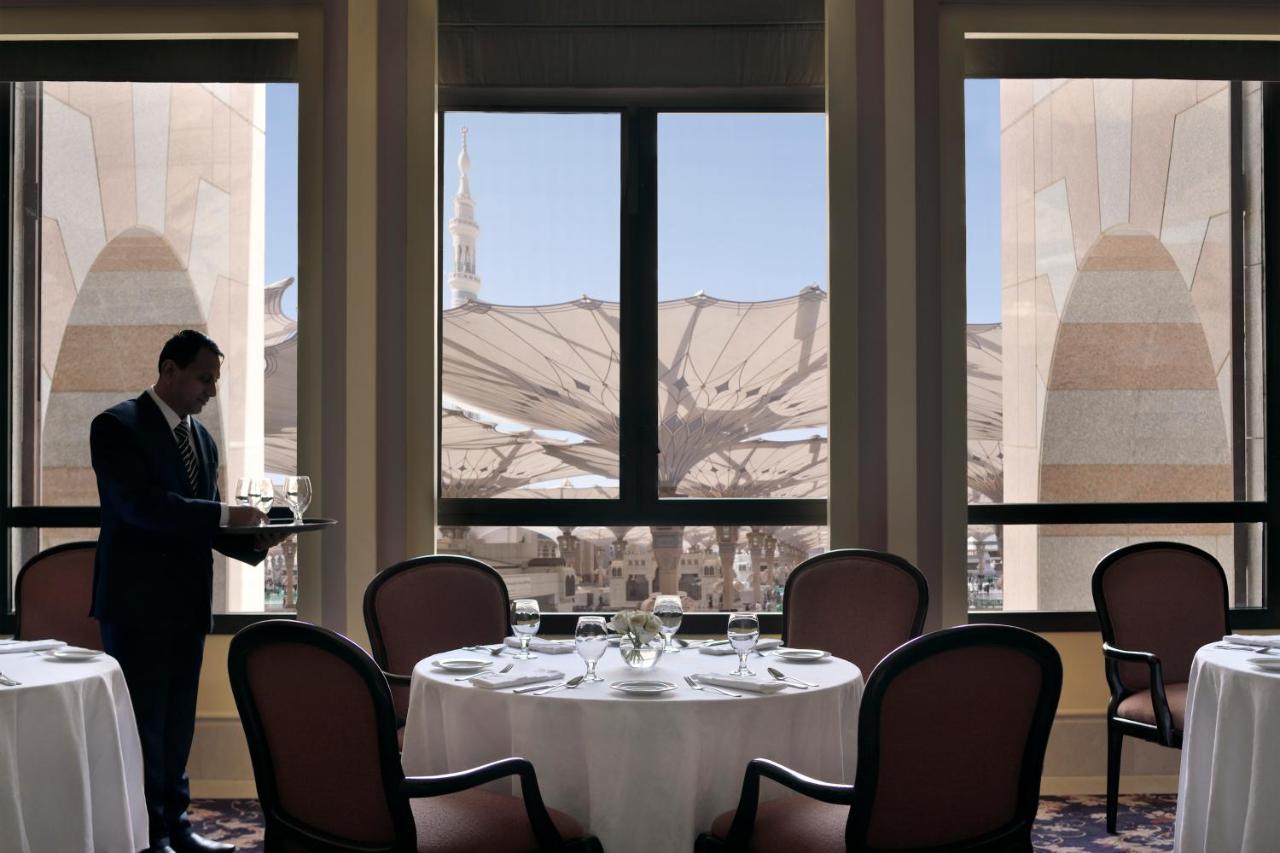 Hotel Intercontinental Dar Al Iman Madinah By Ihg Medina Exterior foto The photo shows a dining area with elegant decor. A waiter is standing to the side, holding a tray. The tables are neatly set with white tablecloths and fine dining arrangements. Large windows provide a view of an outdoor area featuring architectural