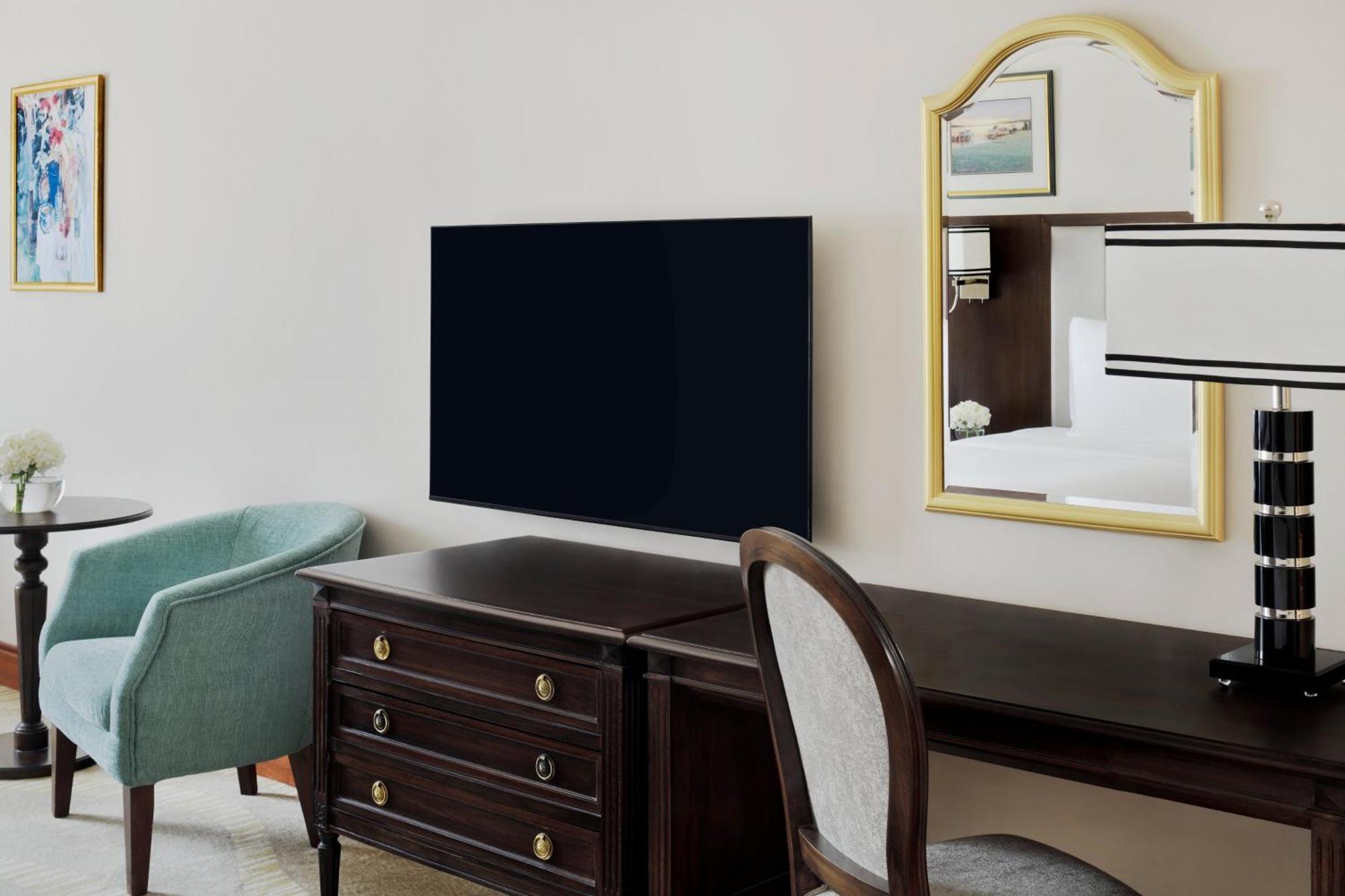 Hotel Intercontinental Dar Al Iman Madinah By Ihg Medina Exterior foto The photo shows a hotel room interior featuring a mounted flat-screen television on the wall. Below the TV, there is a dark wooden dresser with drawers. To the right, there's a wooden desk and a chair with a light-colored seat. A small table with a l