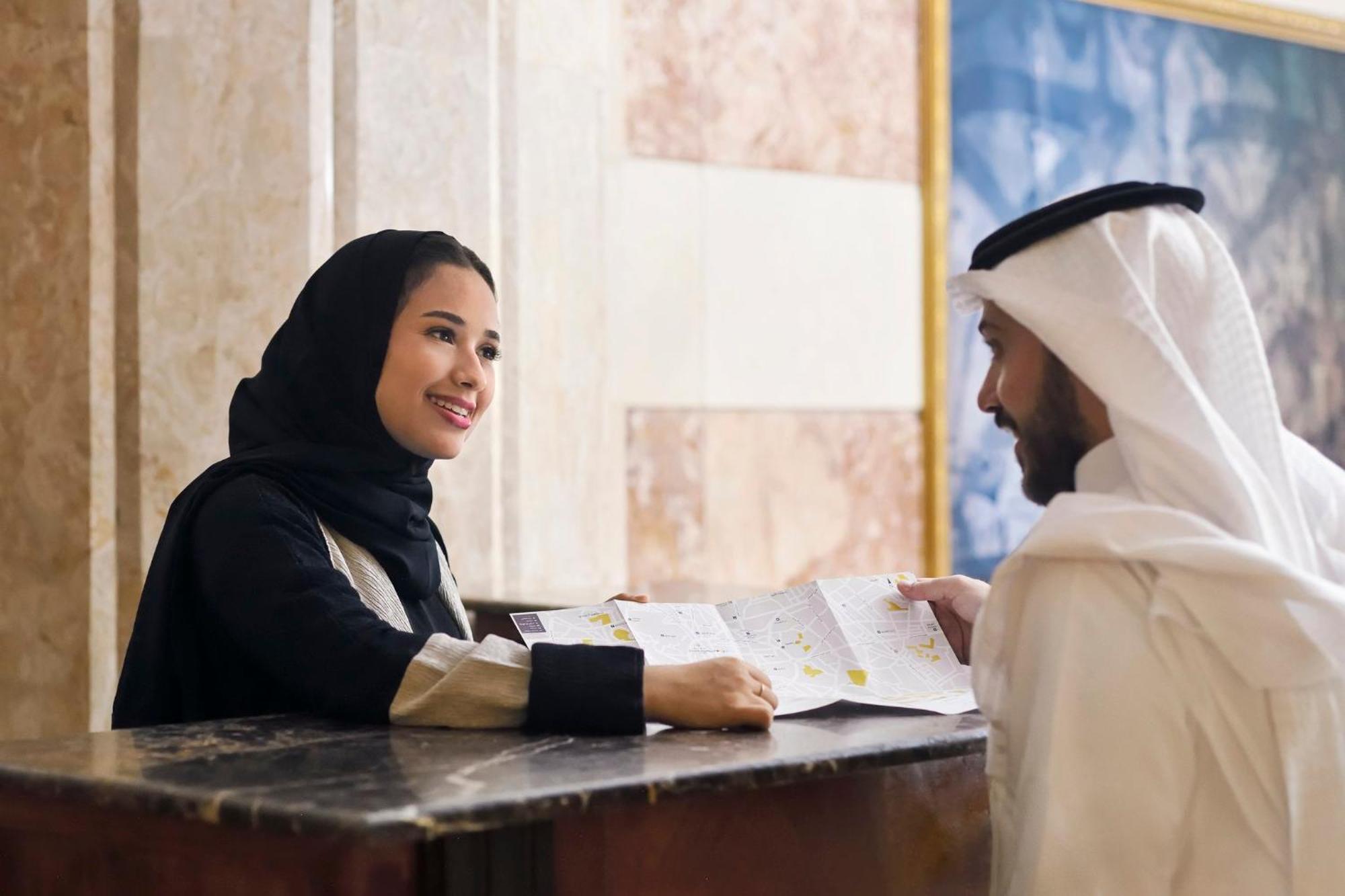 Hotel Intercontinental Dar Al Iman Madinah By Ihg Medina Exterior foto The photo depicts a woman in a black hijab and a light-colored outfit smiling as she interacts with a man wearing traditional Emirati attire, including a white kandura and ghutrah (headscarf). They are at a reception desk in what appears to be a hote