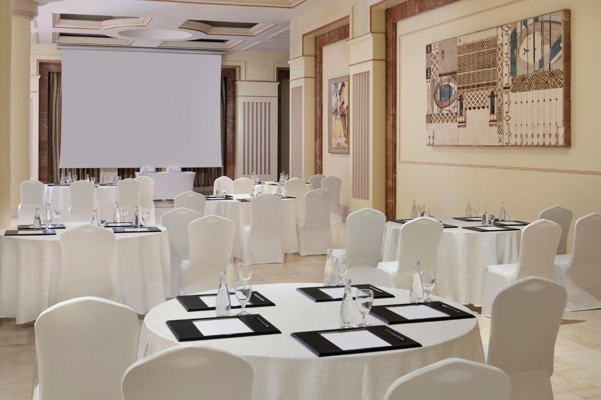 Hotel Intercontinental Dar Al Iman Madinah By Ihg Medina Exterior foto The photo shows a well-decorated event space or meeting room. It features several round tables covered with white tablecloths and surrounded by white chairs. Each table has black placemats and glass water bottles, suggesting a formal setup for a gath