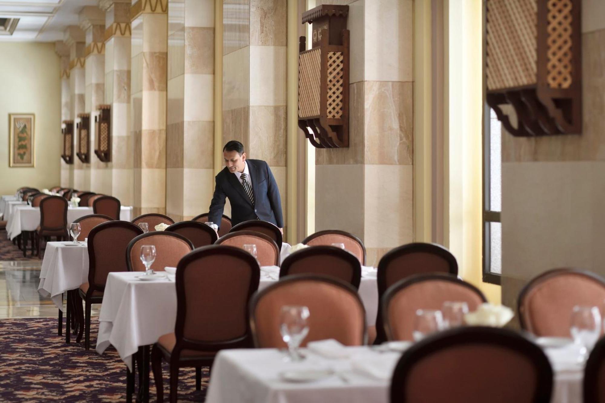 Hotel Intercontinental Dar Al Iman Madinah By Ihg Medina Exterior foto The photo depicts a dining area in an elegant restaurant. There are several tables set with white tablecloths, each accompanied by chairs. A waiter, dressed in a formal suit, is seen arranging or tending to one of the tables. The restaurant features 