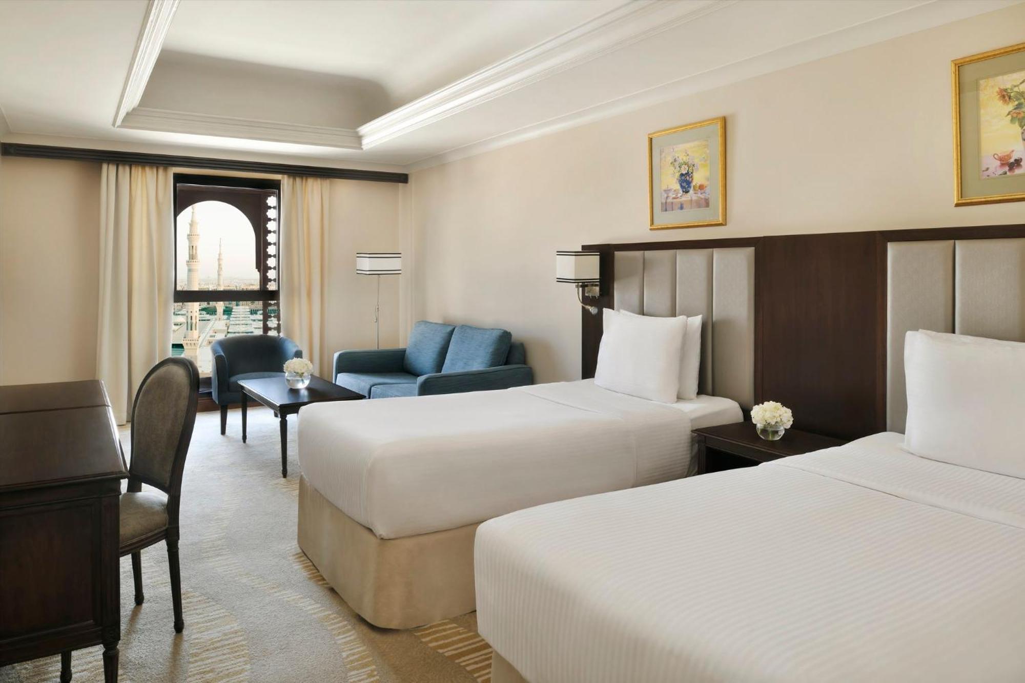 Hotel Intercontinental Dar Al Iman Madinah By Ihg Medina Exterior foto The photo shows a well-appointed hotel room featuring two twin beds with white bedding. There is a small desk with a chair, a comfortable blue sofa, and a coffee table in the sitting area. The room has a warm, neutral color scheme, accented by two pa