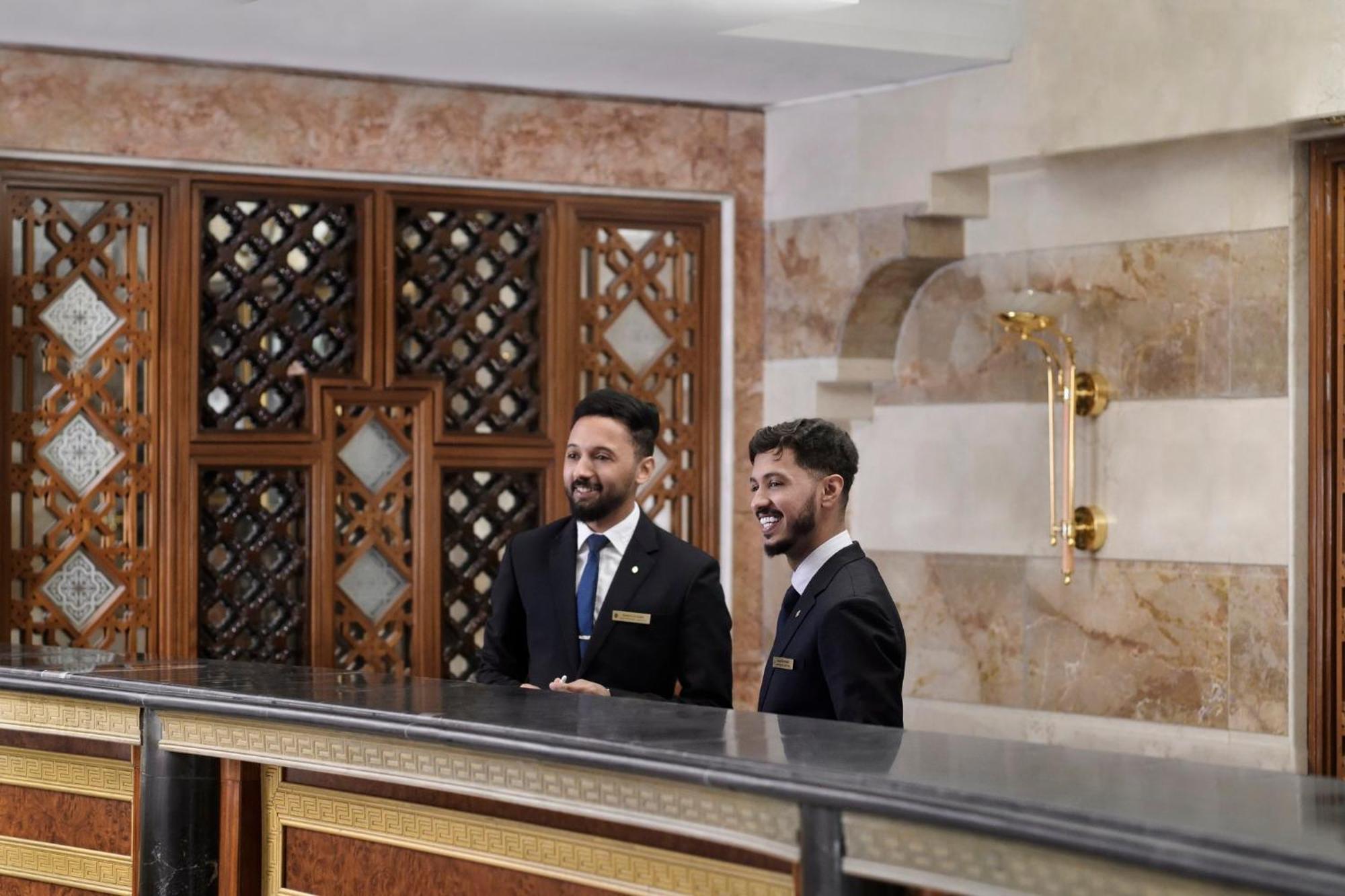 Hotel Intercontinental Dar Al Iman Madinah By Ihg Medina Exterior foto The photo shows two men standing behind a reception desk in an elegant setting, likely a hotel or similar establishment. They are both dressed in formal attire, wearing suits and ties, and appear to be smiling or engaging in conversation. The backgro