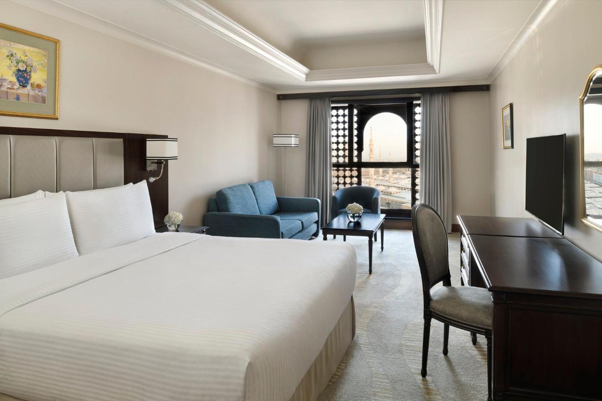 Hotel Intercontinental Dar Al Iman Madinah By Ihg Medina Exterior foto The photo shows a hotel room designed with a modern and comfortable aesthetic. It features a large bed with white linens, a seating area with a blue sofa, and a small table. There is a wooden desk with a chair and a television opposite the bed. Natur