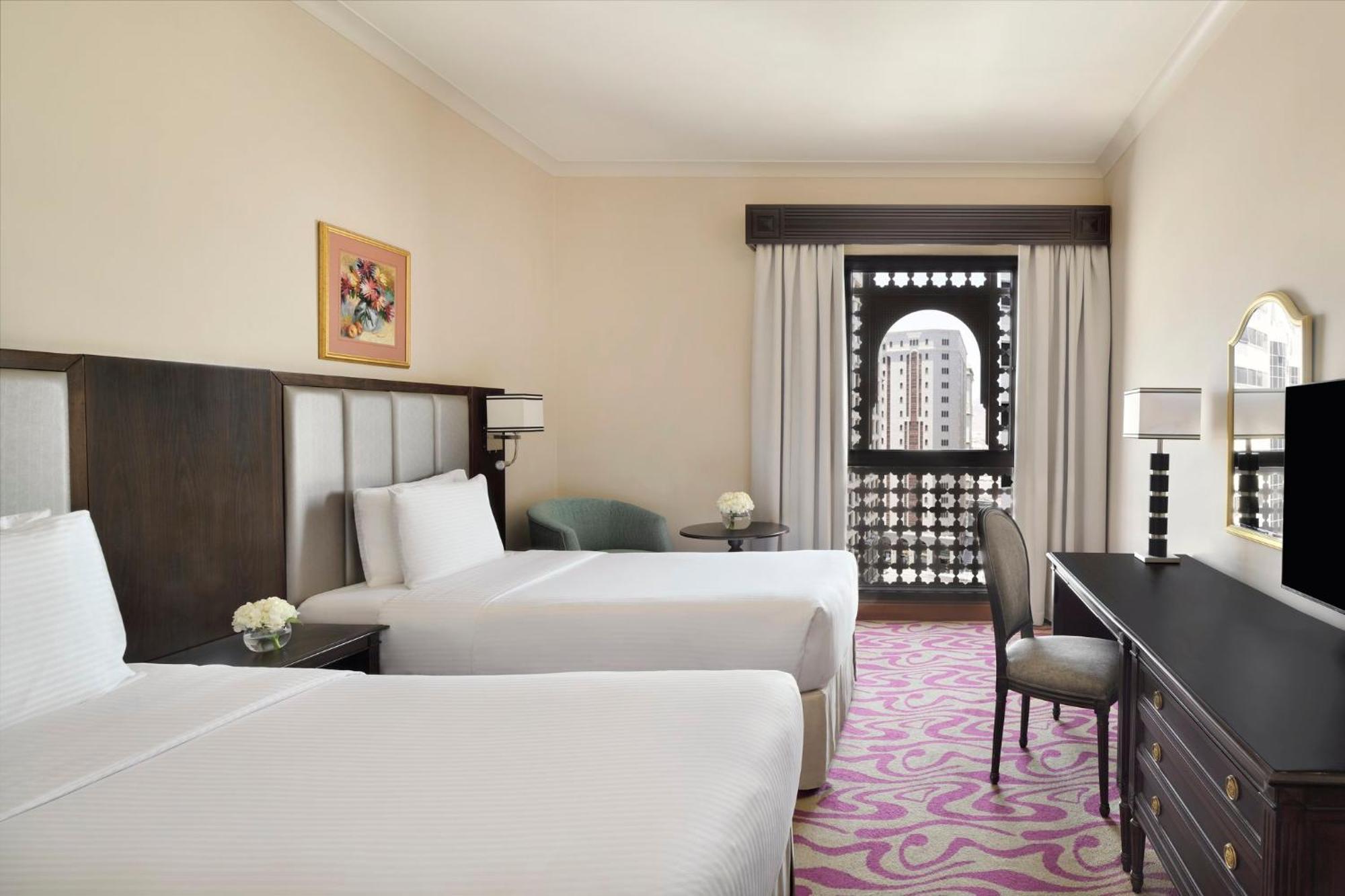 Hotel Intercontinental Dar Al Iman Madinah By Ihg Medina Exterior foto The photo shows a hotel room featuring two twin beds with white bedding. There is a decorative piece of art on the wall above one of the beds. A small round table with a flower arrangement is placed next to the beds. A desk and chair are situated aga