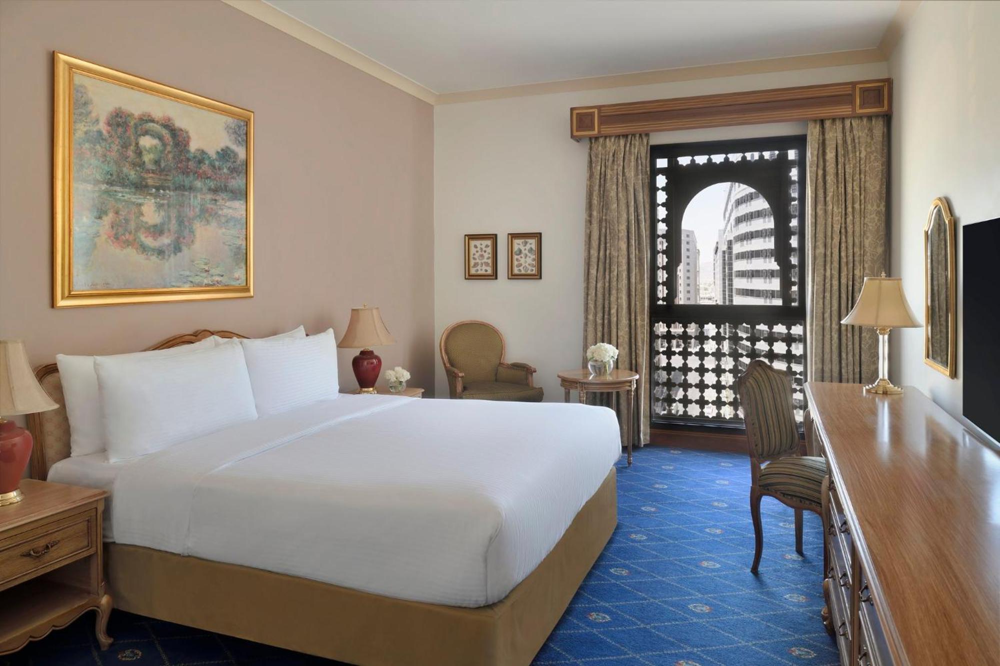 Hotel Intercontinental Dar Al Iman Madinah By Ihg Medina Exterior foto The photo shows a hotel room that features a large, neatly made bed with white linens. There is a framed painting on the wall above the bed. To the side, a small seating area includes a chair and a side table. Opposite the bed, there is a wooden desk