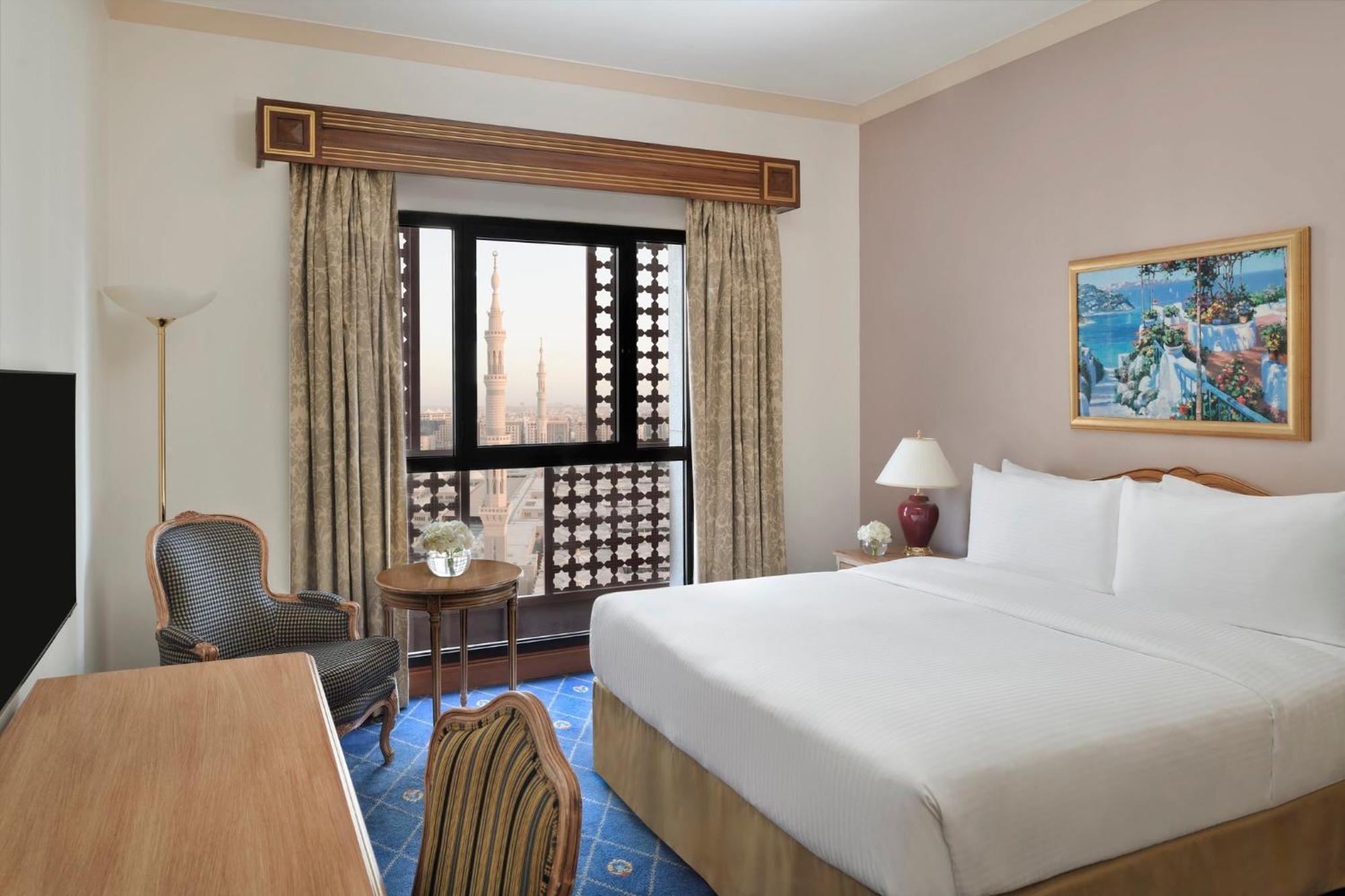 Hotel Intercontinental Dar Al Iman Madinah By Ihg Medina Exterior foto The photo shows a hotel room with a modern design. There is a large bed with white linens placed against the wall. Next to the bed, there is a small bedside table with a lamp. A comfortable armchair is situated nearby, accompanied by a wooden desk. T