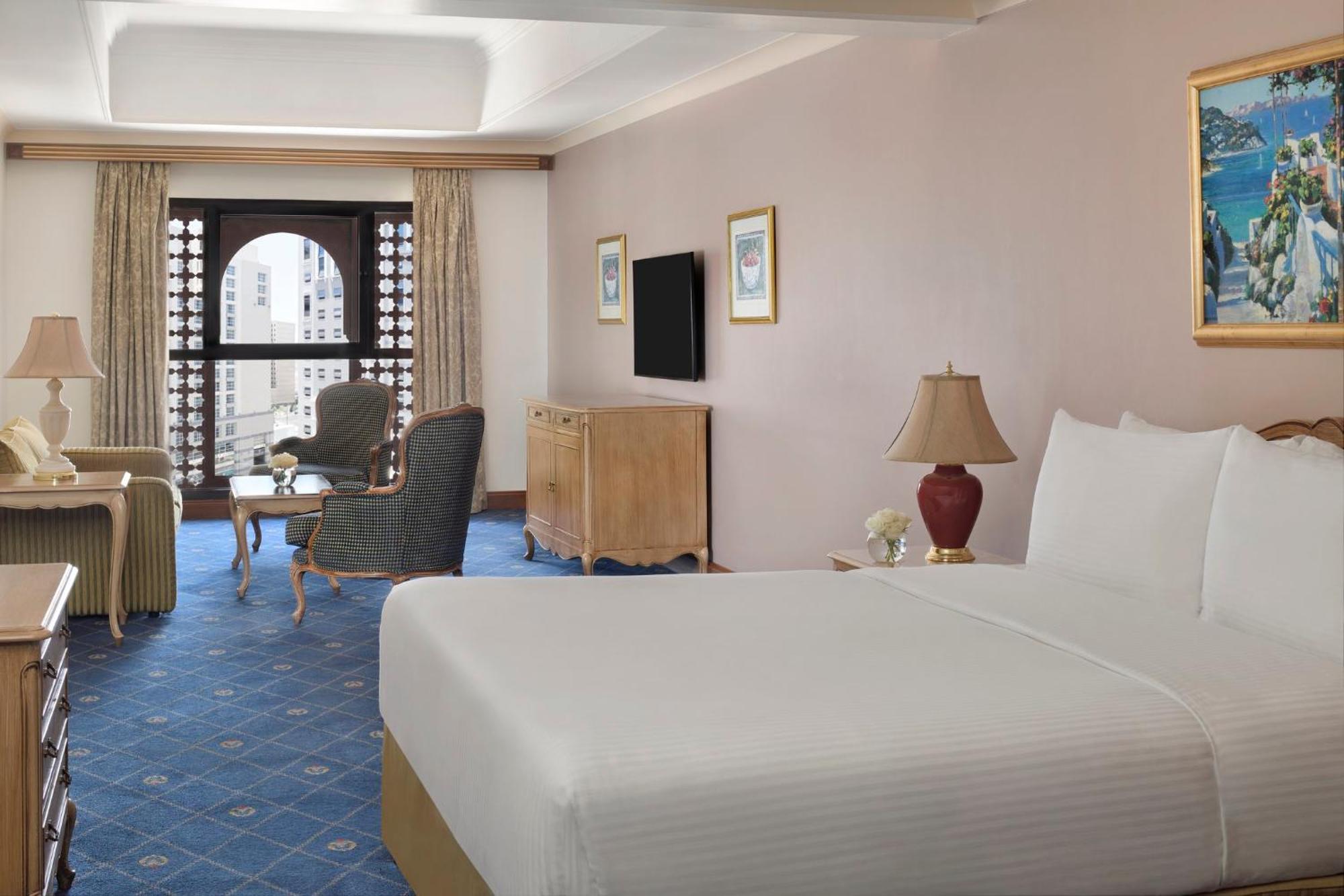 Hotel Intercontinental Dar Al Iman Madinah By Ihg Medina Exterior foto The photo shows a well-furnished hotel room. It features a large bed with a white duvet, two bedside lamps, and decorative paintings on the walls. There is a comfortable seating area with two chairs and a small table. A wooden dresser is positioned a
