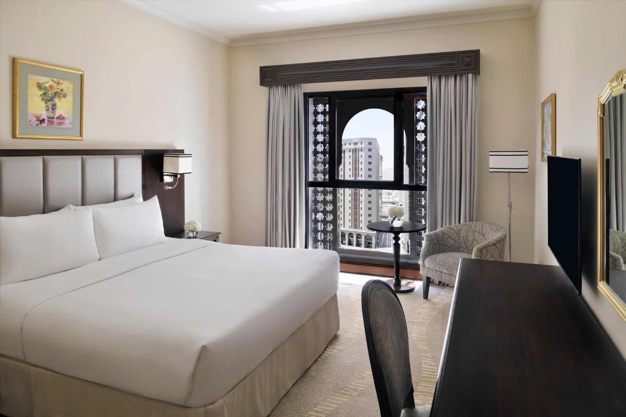 Hotel Intercontinental Dar Al Iman Madinah By Ihg Medina Exterior foto The photo shows a hotel room with a modern and minimalistic design. It features a large bed with white linens, a side table, and a comfortable chair near a window. The window has a view of a cityscape, and elegant curtains frame the window. There is 