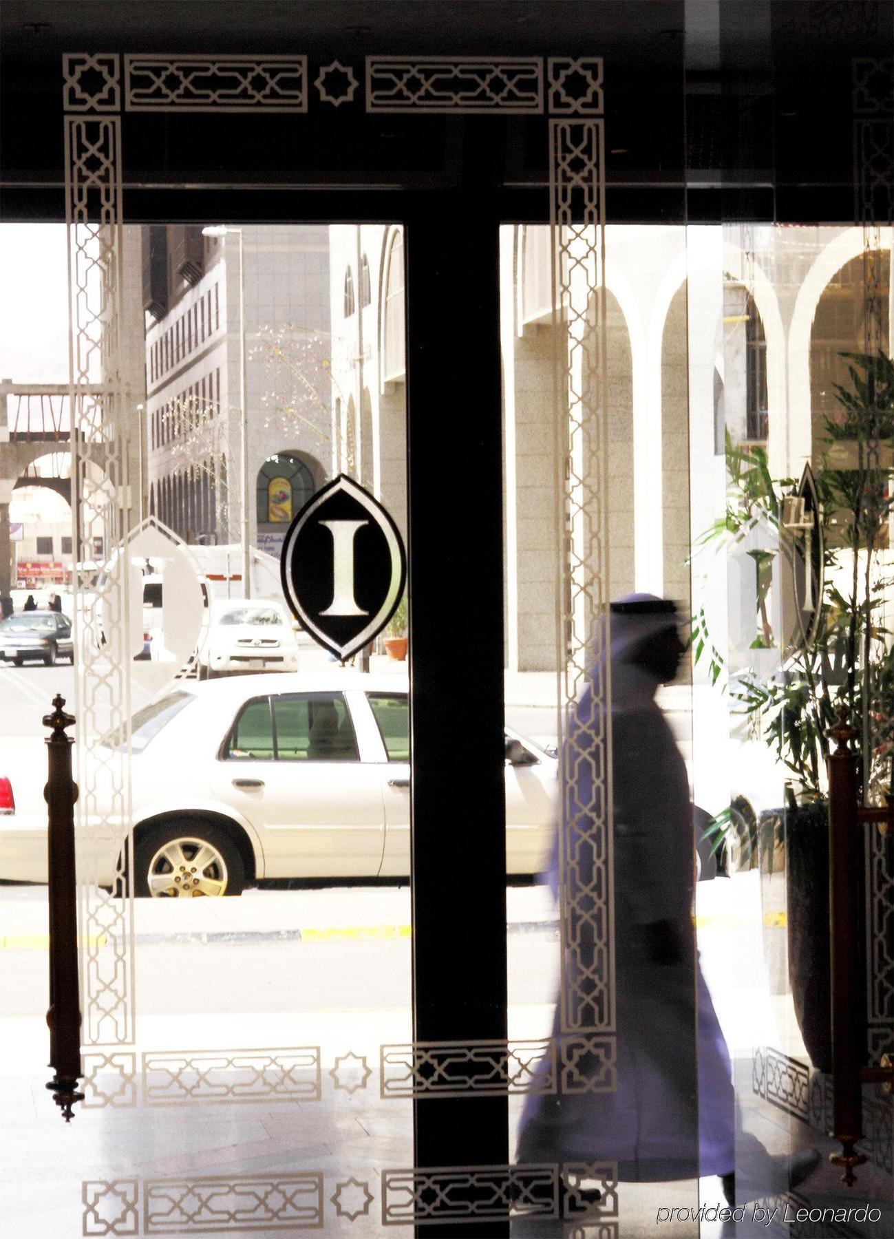 Hotel Intercontinental Dar Al Iman Madinah By Ihg Medina Interior foto The photo shows a view through a glass door or window, featuring an elegant design with a prominent "I" symbol displayed. Through the glass, there are some parked vehicles visible, including a white car. In the foreground, a shadowy figure of a perso