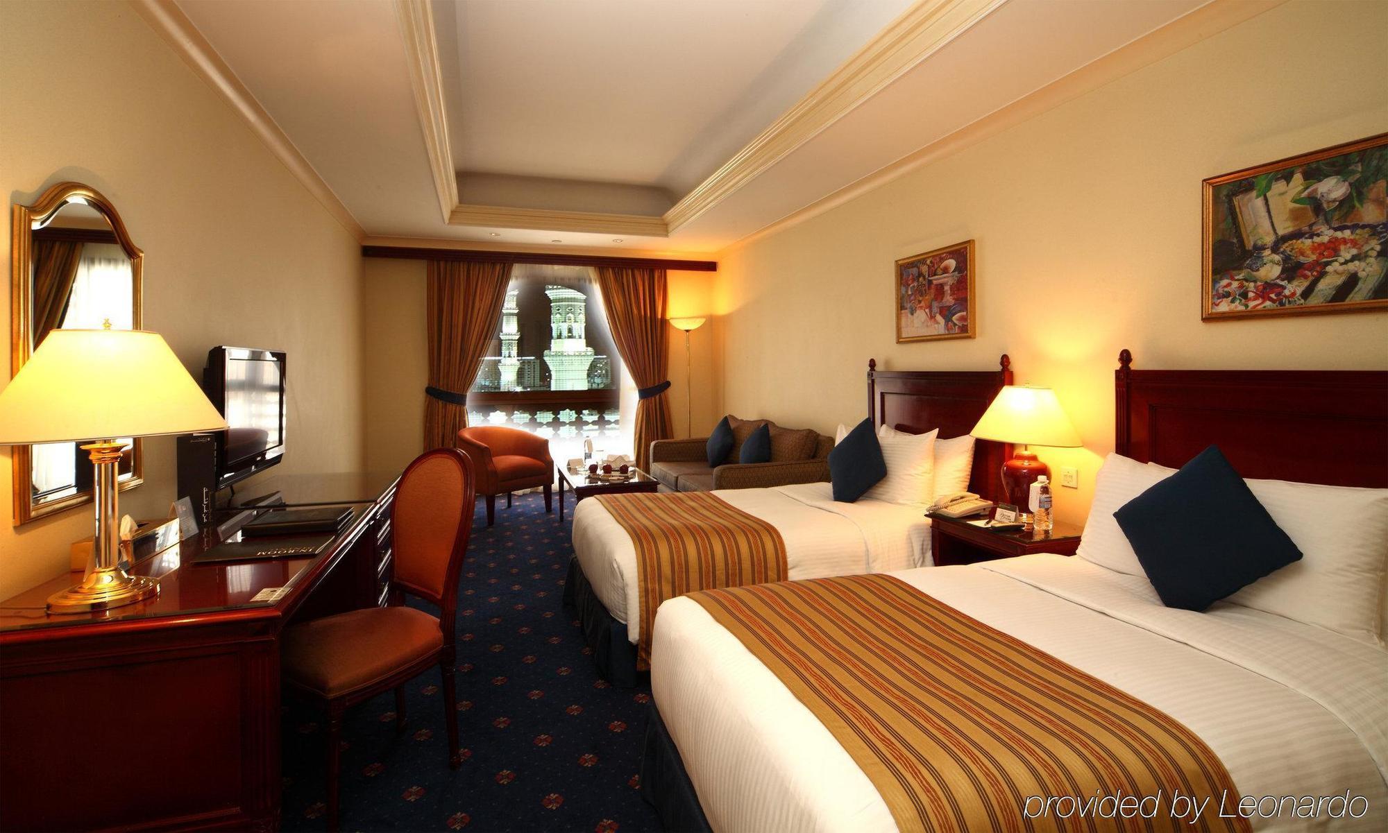 Hotel Intercontinental Dar Al Iman Madinah By Ihg Medina Habitación foto The photo shows a hotel room featuring two beds with white linens and brown striped throws. There is a wooden desk and chair, alongside a television. The walls are painted in a warm tone, and there are decorative paintings. Large windows with curtain