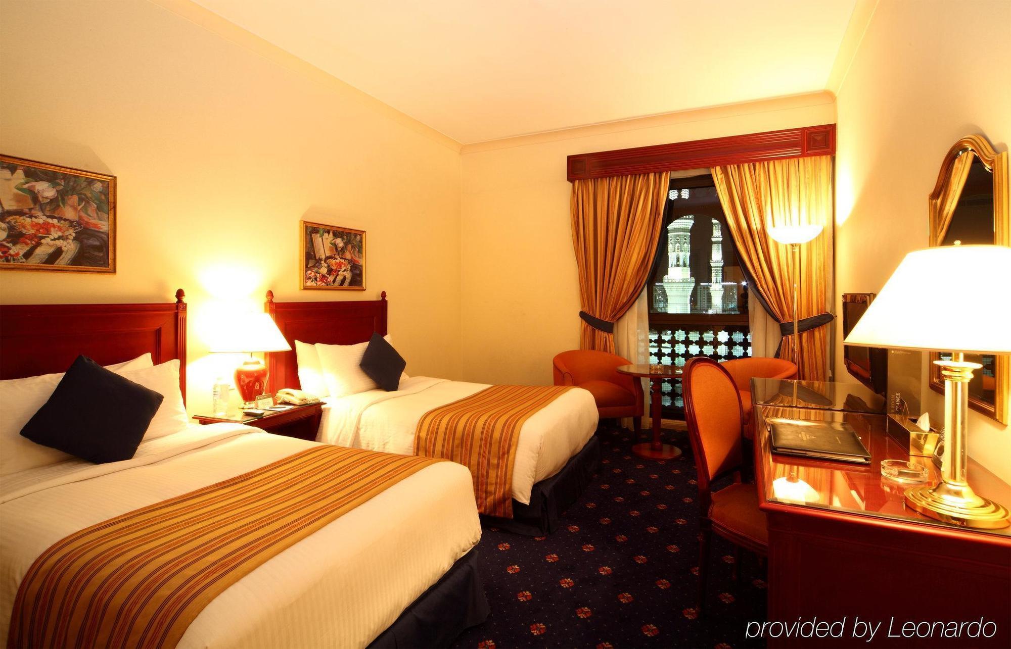 Hotel Intercontinental Dar Al Iman Madinah By Ihg Medina Habitación foto The photo shows a hotel room featuring two twin beds with white linens and brown striped bedspreads. There are decorative pillows on the beds, and the room has warm-colored walls. To the side, there's a small sitting area with a round table and a cha