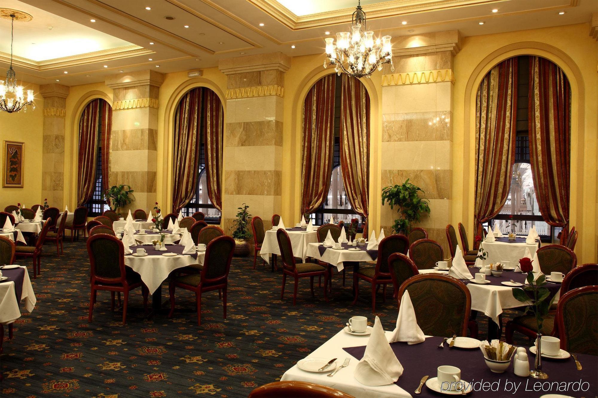 Hotel Intercontinental Dar Al Iman Madinah By Ihg Medina Restaurante foto The photo depicts an elegant restaurant interior. The space features ornate decor with tall windows draped in rich, patterned curtains. Tables are neatly set with white tablecloths and napkins, and there are plush chairs arranged around them. The flo