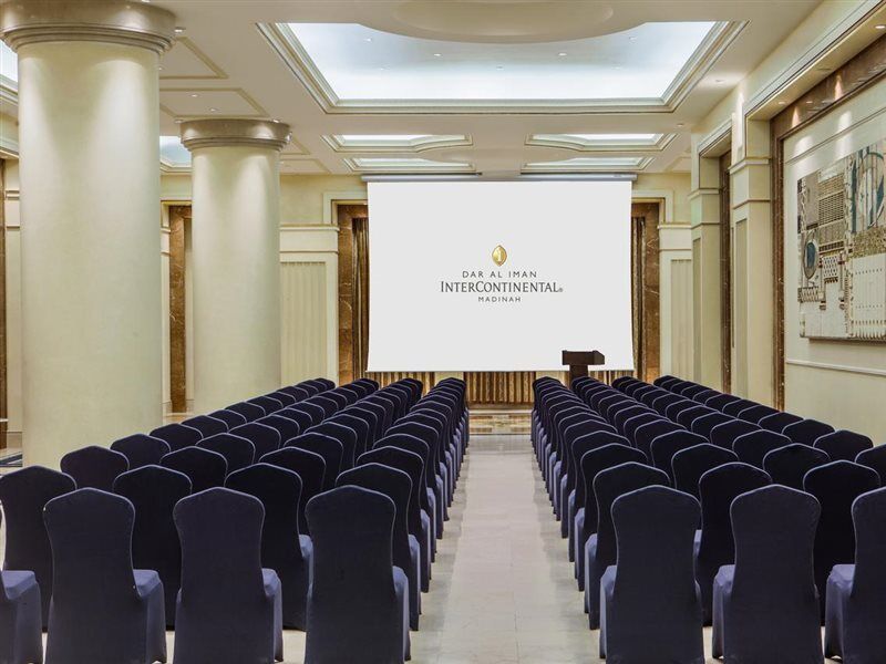 Hotel Intercontinental Dar Al Iman Madinah By Ihg Medina Exterior foto The photo shows a conference or meeting room set up. There are rows of dark blue chairs arranged facing a large screen at the front of the room. The screen displays the logo of the Intercontinental hotel, indicating that this space is likely intended