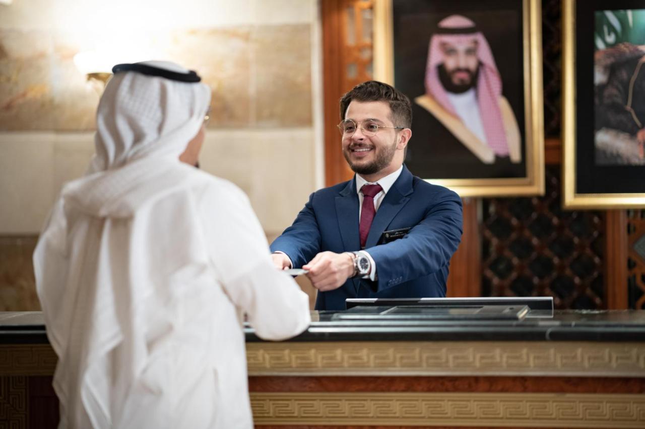Hotel Intercontinental Dar Al Iman Madinah By Ihg Medina Exterior foto The photo shows a hotel reception area. A male receptionist, dressed in a navy suit with a tie, is smiling and handing an item to a guest who is wearing traditional white attire, possibly a kandura or thobe. The background features elegantly decorate
